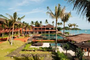 A tropical beachfront resort with lush palm trees, wooden buildings, a large outdoor pool, and views of the ocean with boats in the distance under a clear blue sky.