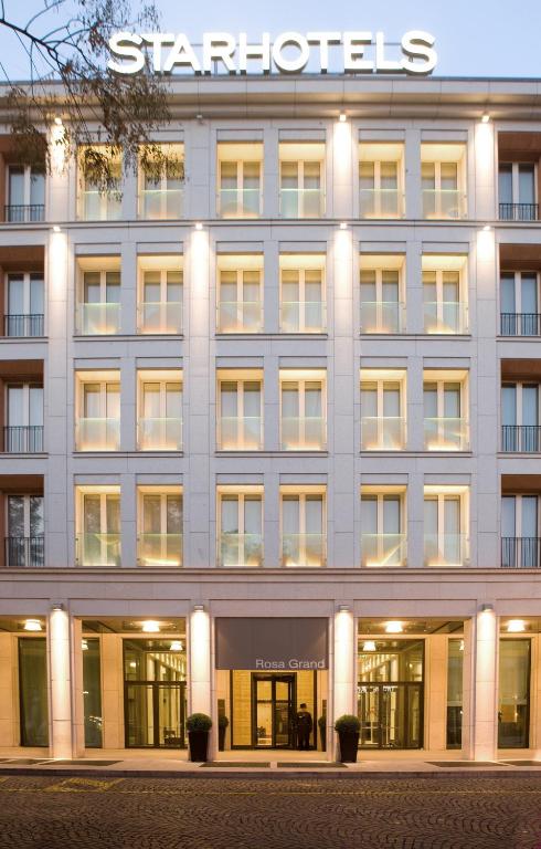 The exterior of a modern hotel building with large windows and illuminated lights at dusk. The sign STARHOTELS is displayed on the roof, and Rosa Grand is above the entrance.