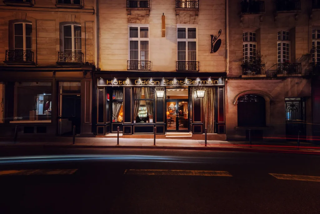 A warmly lit entrance to the Hôtel Odéon Saint-Germain at night, with elegant curtains, glowing signs, and light trails from passing cars on the quiet street in front.