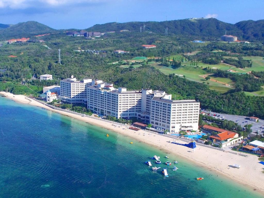 Aerial view of a large beachfront resort with white buildings beside a sandy beach and clear blue-green water, surrounded by lush green hills and a golf course in the background.