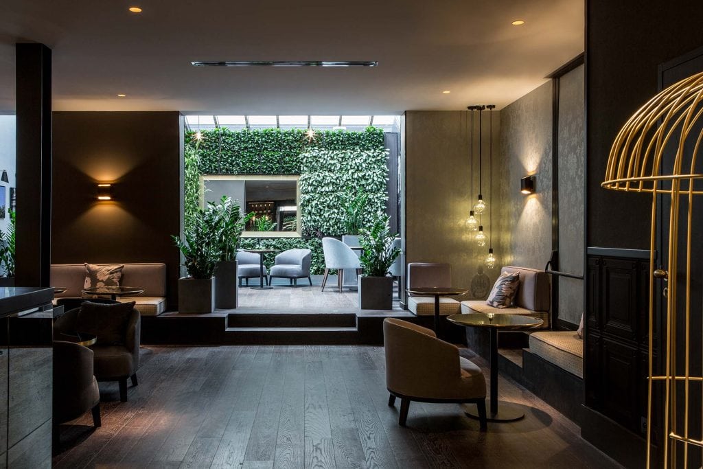 Modern lounge with soft lighting, armchairs, small tables, and lush potted plants. In the background, a bright patio with greenery, white chairs, and a green living wall is visible through an open glass wall.