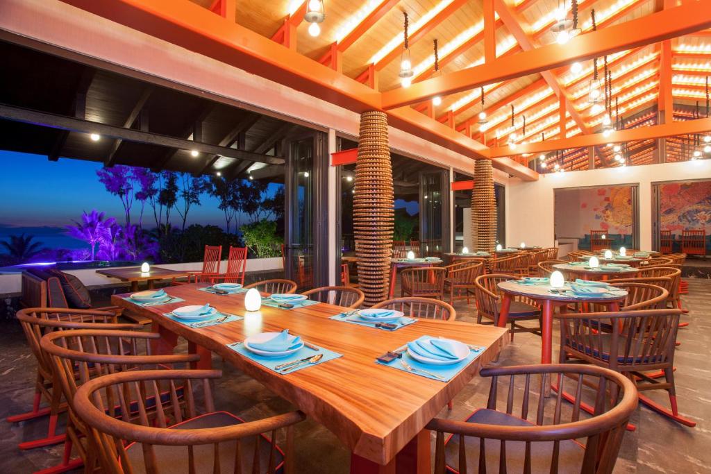 Modern restaurant interior with wooden tables set with blue napkins and plates, wooden chairs, warm lighting, and large windows overlooking trees and a nighttime outdoor view with purple lighting.