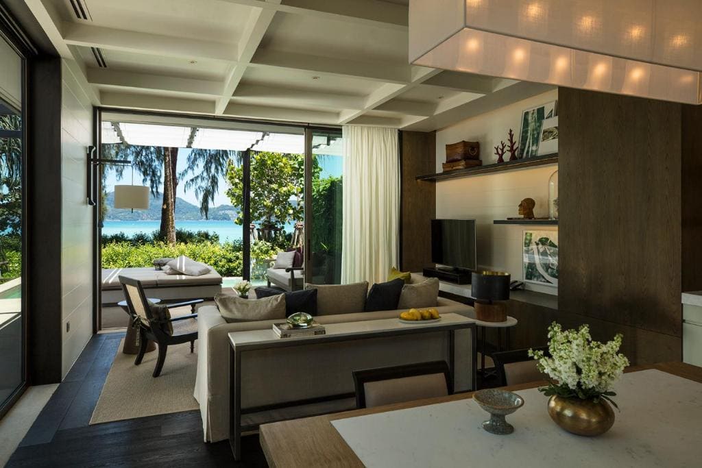 A modern living room with neutral colors, large windows, and a scenic view of lush greenery and water outside. The space includes a sofa, chairs, TV, open shelves, and a dining table with flowers.