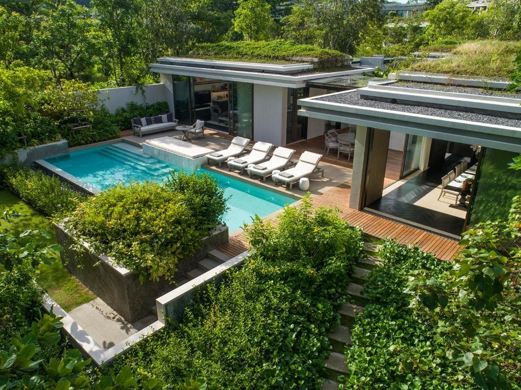 A modern villa with a private rectangular pool, sun loungers, and shaded outdoor seating, surrounded by lush greenery and landscaped gardens. Large glass doors connect the indoor and outdoor spaces.