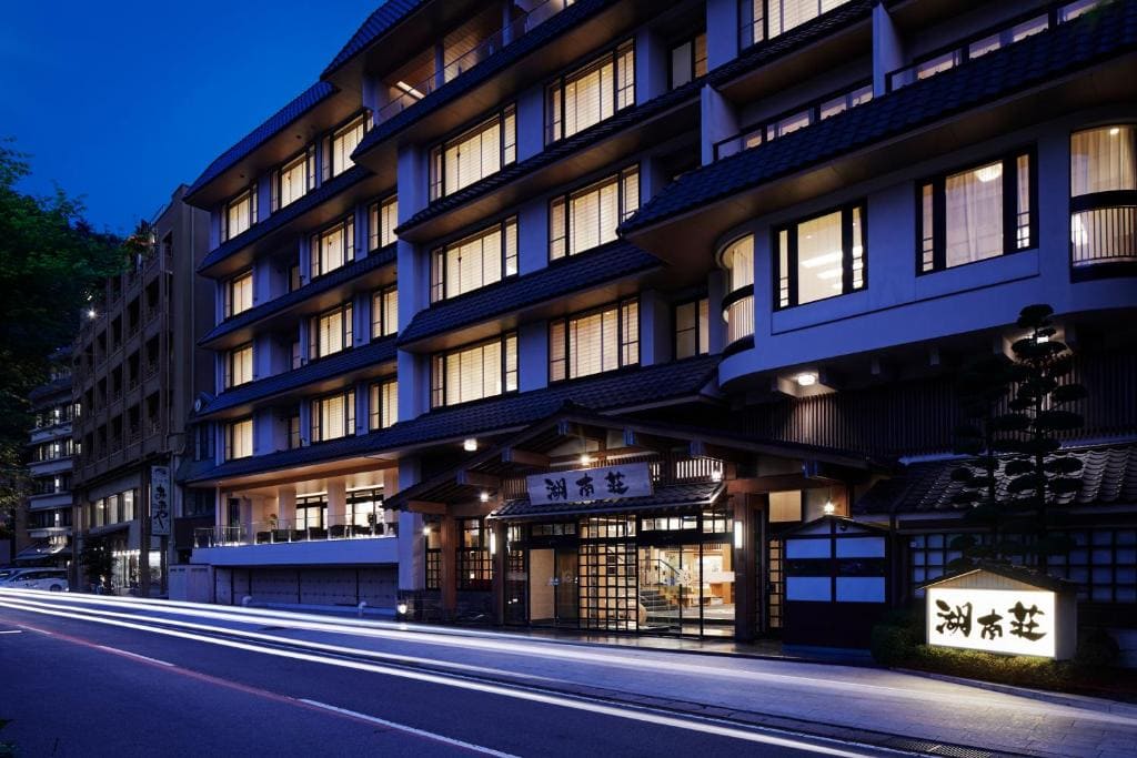 A multi-story traditional Japanese inn, warmly lit at dusk, stands alongside a street with light trails from passing cars, creating a serene and inviting nighttime atmosphere.