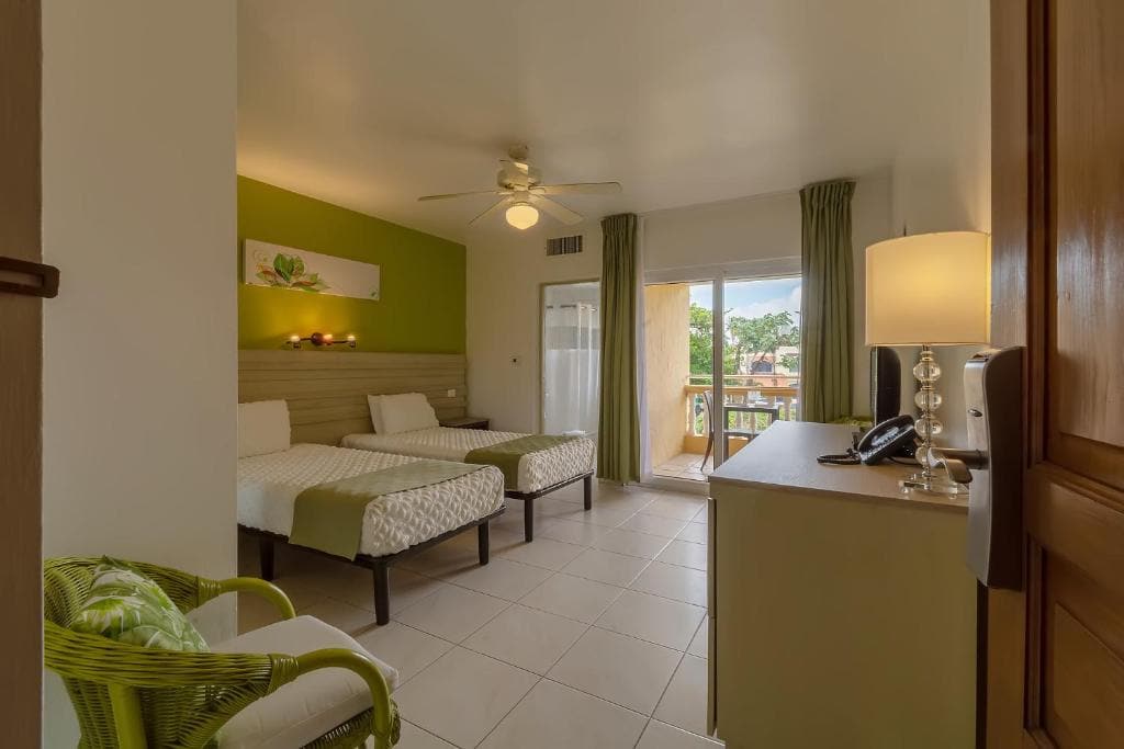 A bright hotel room with two single beds, green accent wall, a ceiling fan, a desk with a lamp and phone, a green armchair, and a balcony with curtains letting in natural light.
