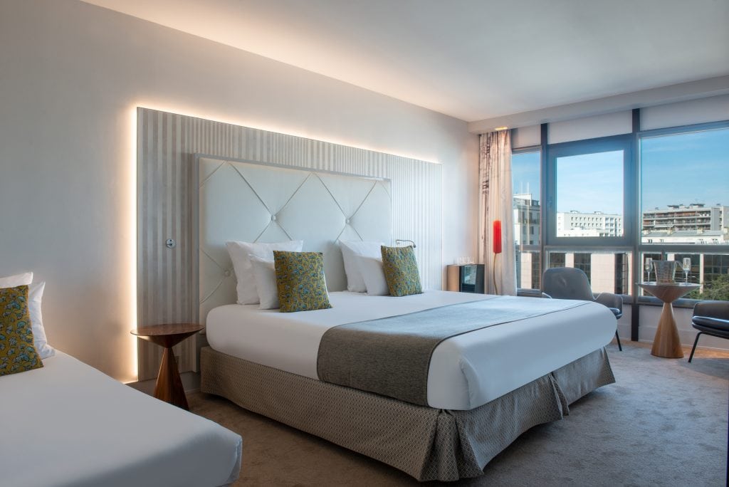 A modern hotel room features a large bed with crisp white linens, gray accents, and decorative pillows. There is ample natural light from large windows, contemporary furnishings, and a city view outside.