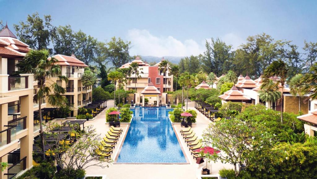 A luxurious resort features a long central swimming pool surrounded by lounge chairs, tropical trees, and multi-story buildings with red-tiled roofs, set against a backdrop of lush greenery and distant hills.