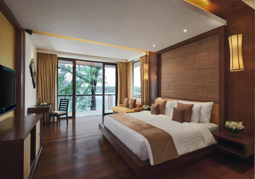 A spacious hotel room with a large bed, brown and white bedding, wooden floors, a desk and chair by floor-to-ceiling windows, and a view of trees and water outside. Warm lighting and modern decor create a cozy atmosphere.