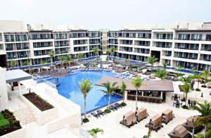 Modern resort with multiple stories overlooking a large, irregularly-shaped outdoor pool, surrounded by loungers, palm trees, poolside seating areas, and cabanas; clear sky above.