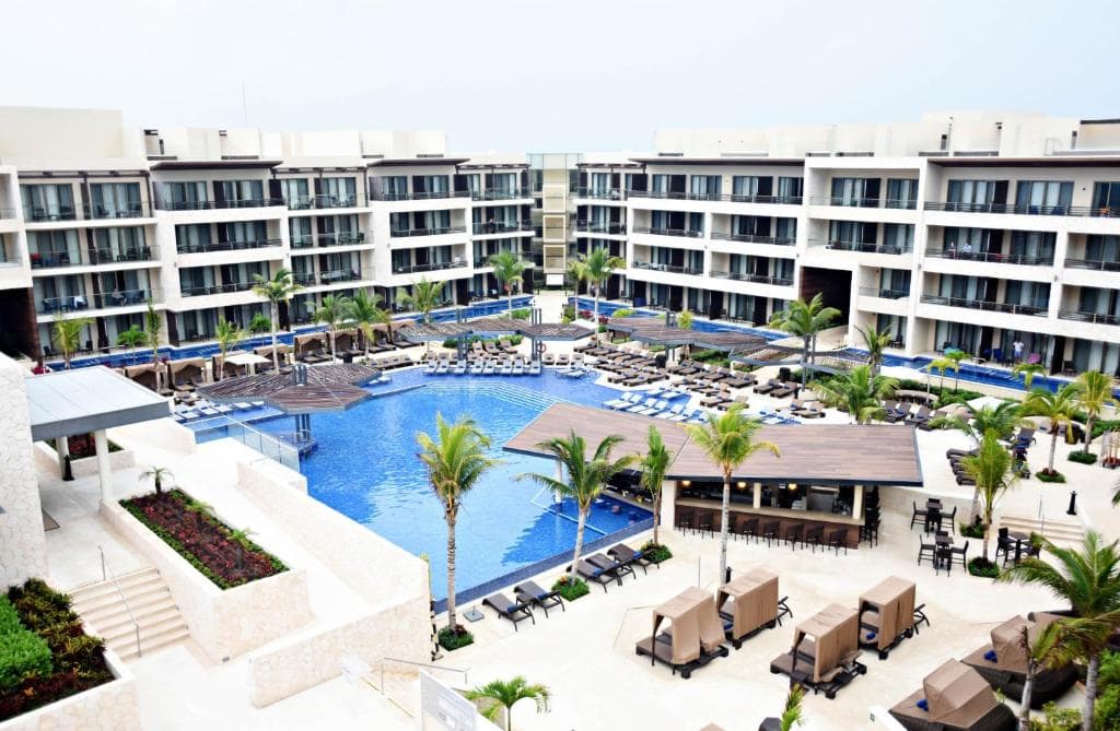 Modern resort with multiple stories overlooking a large, irregularly-shaped outdoor pool, surrounded by loungers, palm trees, poolside seating areas, and cabanas; clear sky above.
