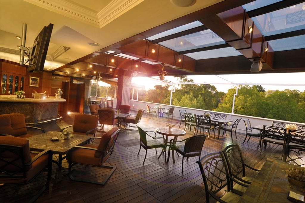 Spacious outdoor restaurant patio with various tables and chairs, warm lighting, a bar area, and large windows offering a view of trees at sunset.
