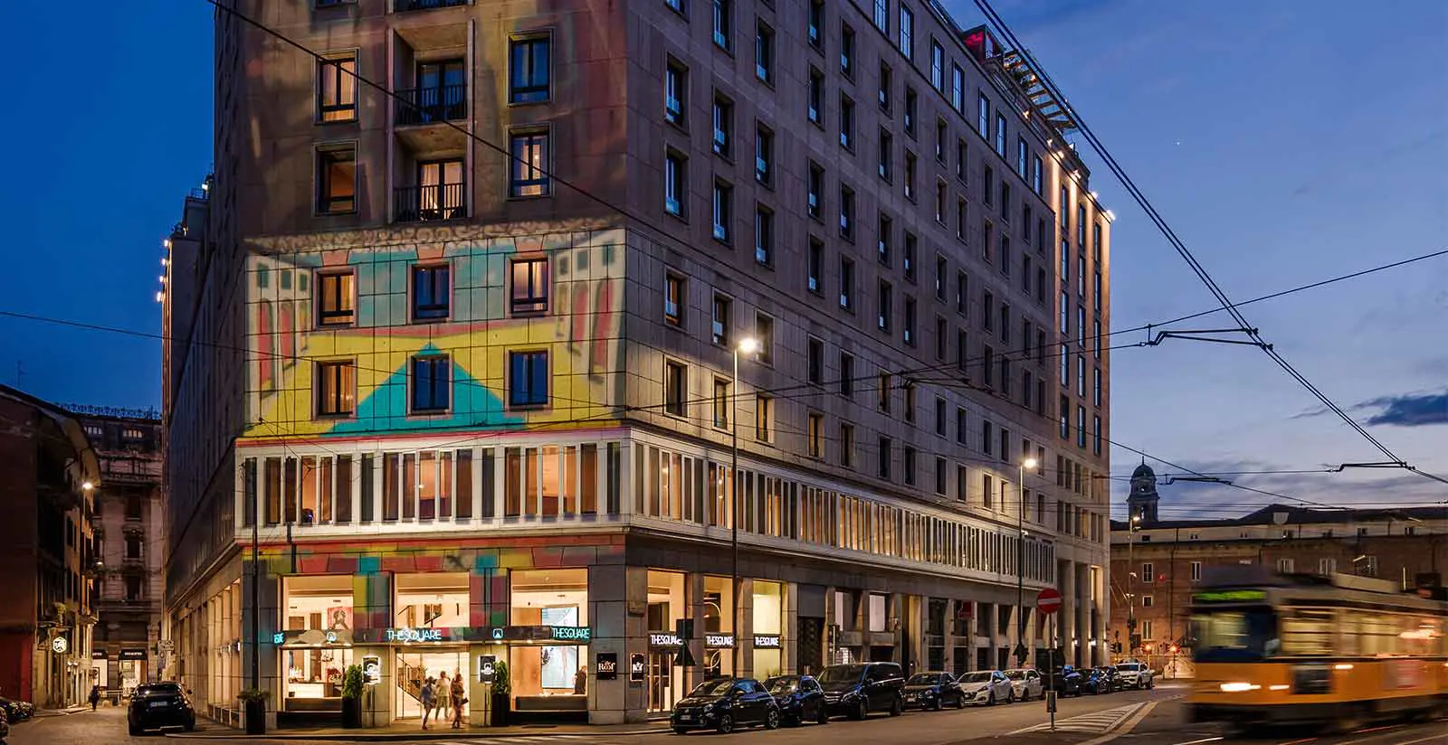 A large, modern building with colorful geometric patterns on its facade is illuminated at dusk. Cars are parked along the street, and a yellow tram is passing by on the right. The sky is deep blue.