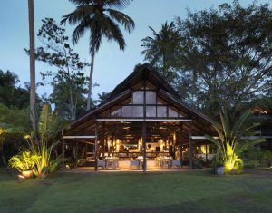 Open-air restaurant with a high, peaked wooden roof surrounded by lush tropical plants and palm trees, warmly lit inside during dusk, with tables and chairs set for dining.