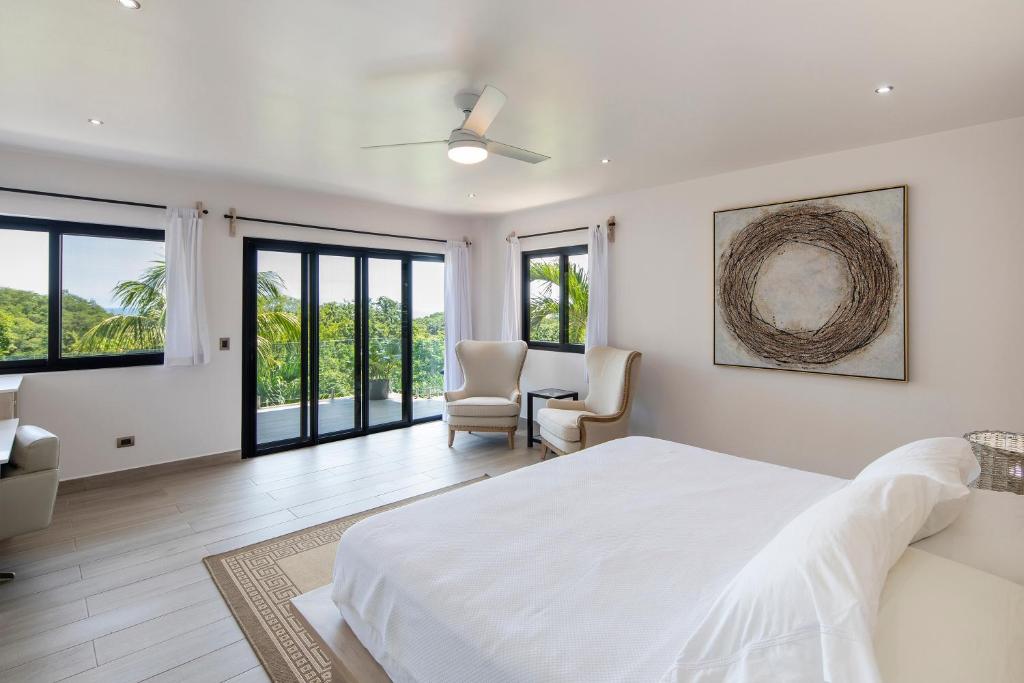 A bright, modern bedroom with large windows and glass doors, a white bed, two armchairs near the corner, a ceiling fan, minimal decor, and views of lush greenery outside.