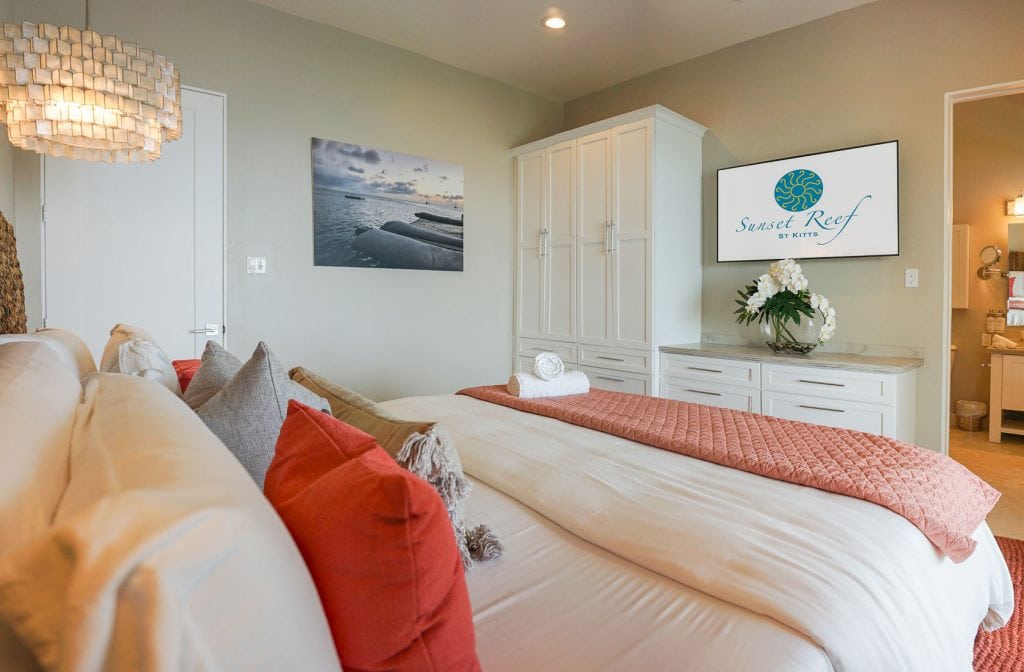 A cozy bedroom with a neatly made bed featuring white and coral pillows and a coral throw. A large TV displays “Sunset Reef.” There are built-in white wardrobes, a seascape photo on the wall, and flowers on a dresser.