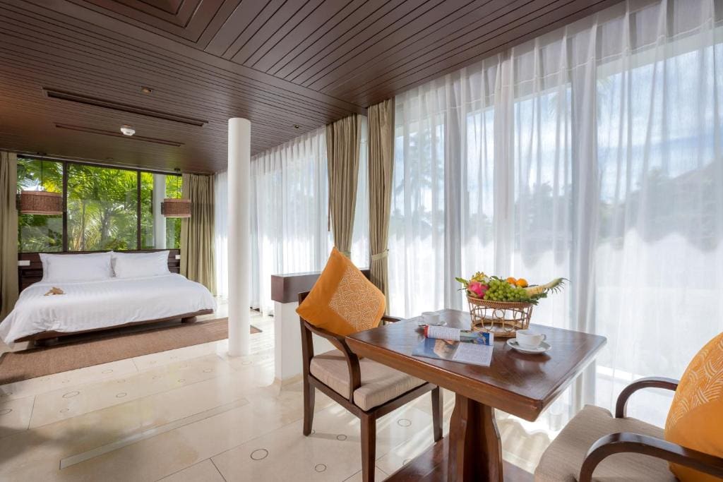 A bright, modern hotel room with a large bed, wooden table and chairs, fruit basket, and floor-to-ceiling windows with sheer curtains letting in natural light and views of greenery outside.