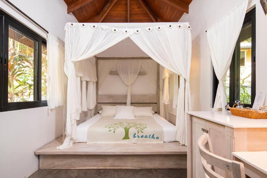 A cozy bedroom with a canopy bed draped in white curtains, white bedding, and a blanket featuring a tree and the word breathe. Sunlit windows with white curtains and a wooden desk with a chair are also visible.