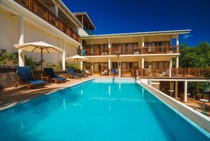 A clear blue swimming pool with lounge chairs and umbrellas sits in front of a two-story hotel with wooden balconies on a sunny day.