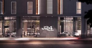 Modern hotel exterior at night featuring large glass windows, sleek dark facade, and illuminated vertical light accents. The sign reads J24 Hotel Milano in white letters. Reflections of cars and trees are visible in the glass.