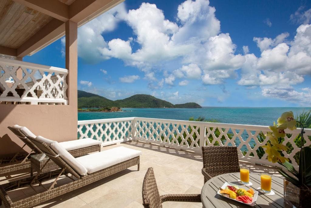 A sunny terrace with lounge chairs, a round table set with fruit and orange juice, and a view of the ocean, green hills, and a blue sky with fluffy clouds.