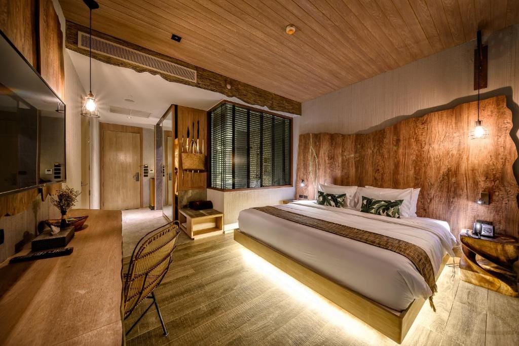 Modern hotel room with wooden accents, a king-sized bed with white linens and decorative pillows, a desk with chair, pendant lights, large TV, and a glass-walled bathroom. Warm, ambient lighting creates a cozy atmosphere.