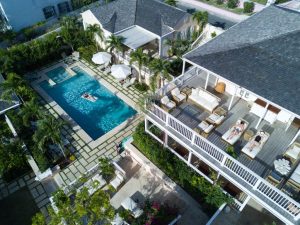 Aerial view of a luxurious villa with a rectangular swimming pool, people relaxing on pool floats, sun loungers and shaded seating areas, surrounded by palm trees and greenery.