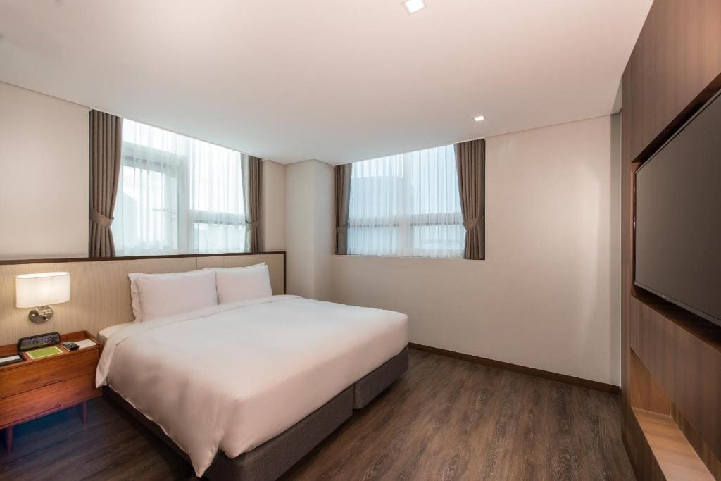 A modern hotel room with a large bed, white bedding, wooden floors, two curtained windows, a bedside table with a lamp, and a wall-mounted TV. The room is well-lit and neatly arranged.