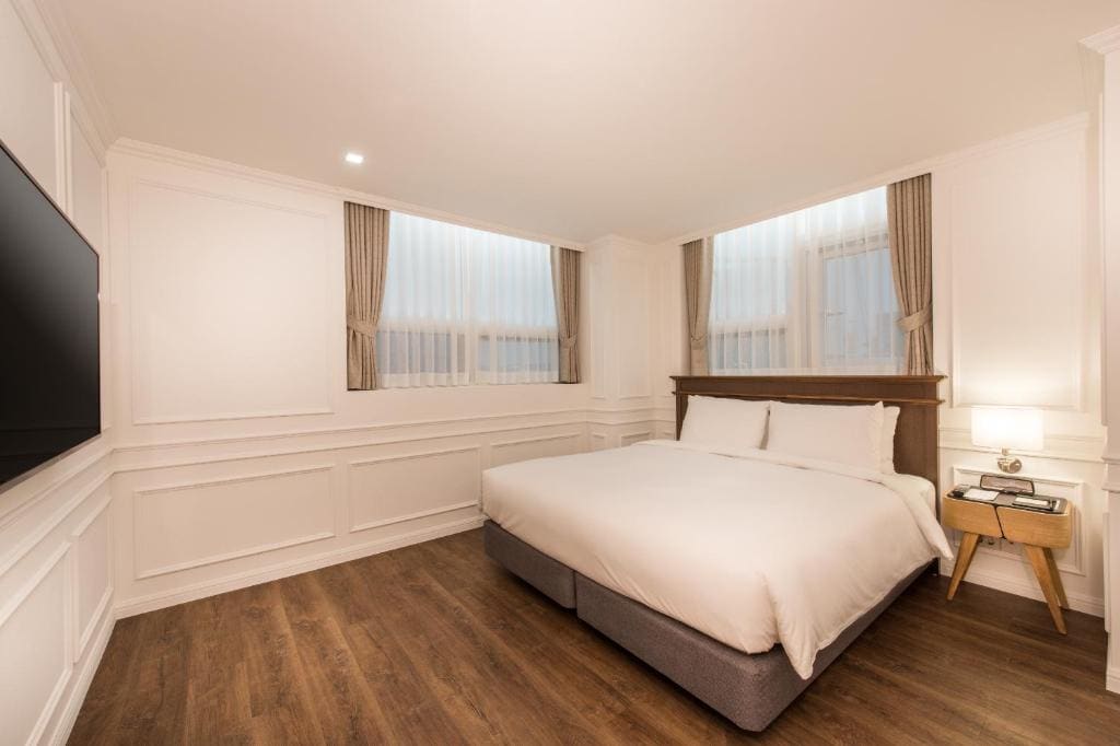 A bright, modern hotel room with a large bed, white bedding, wood floors, a bedside table with a lamp, two curtained windows, white paneled walls, and a flat-screen TV mounted on the wall.