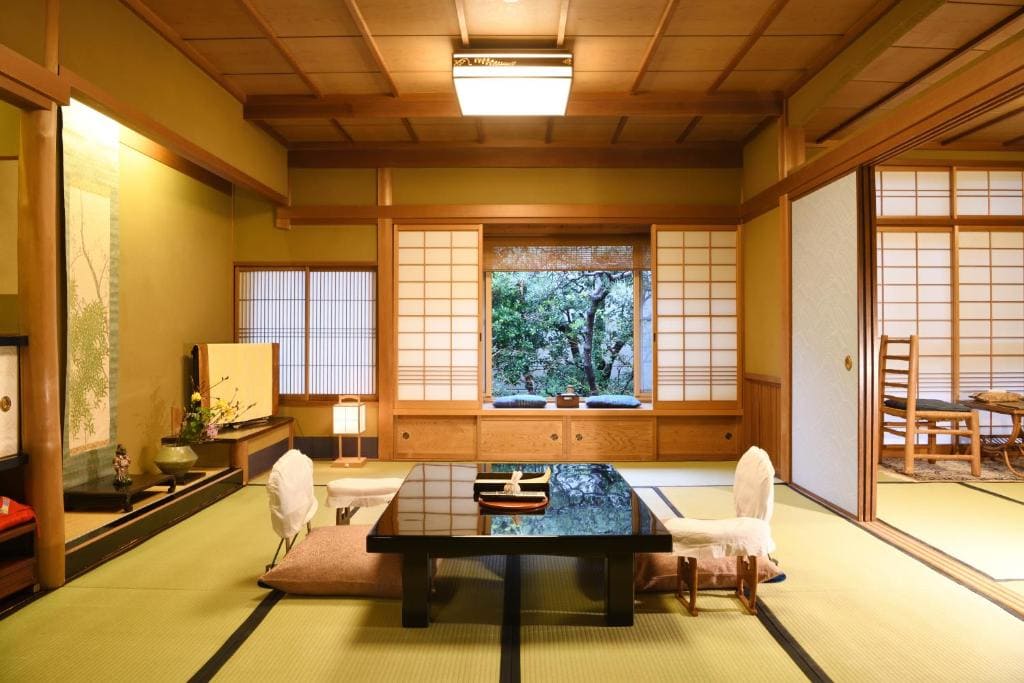 A traditional Japanese tatami room with low table, floor cushions, shoji screens, and a garden view through the window. Minimalist décor with warm lighting and wooden accents.