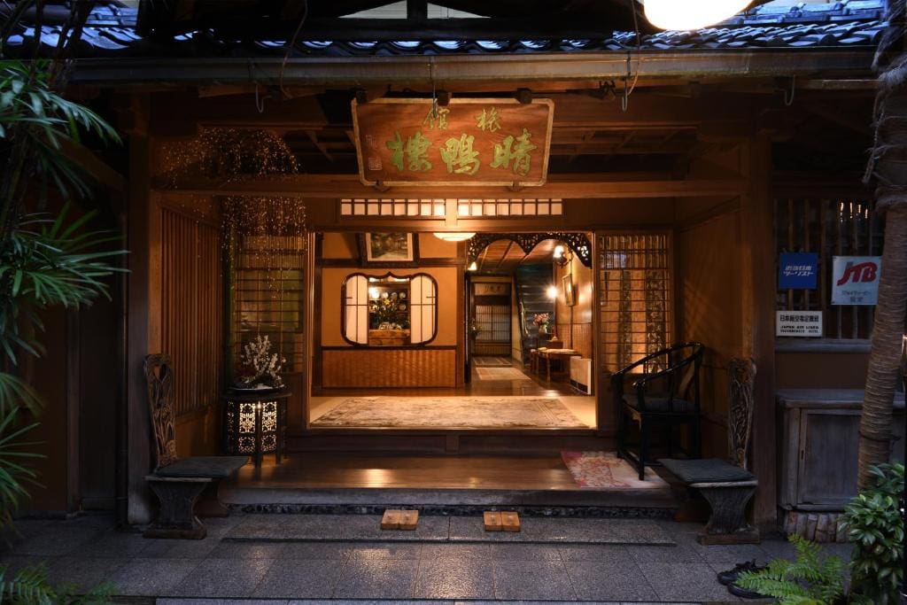 Warmly lit entrance to a traditional Japanese inn with sliding wooden doors, tatami mats, decorative plants, and wooden signboard above, creating a welcoming and serene atmosphere.