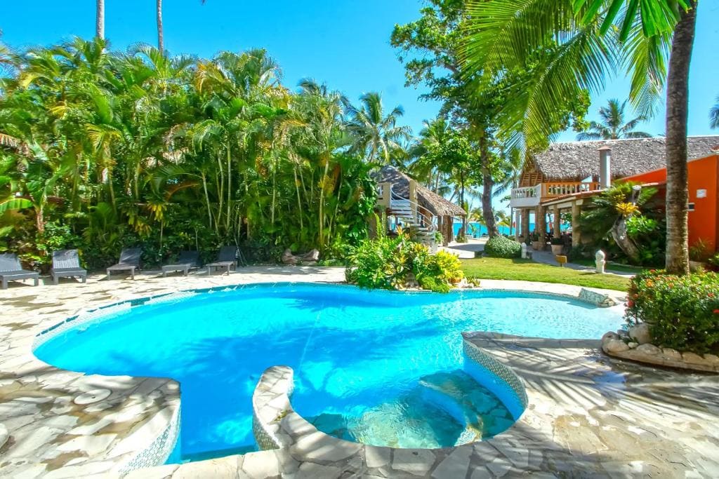 A tropical outdoor pool surrounded by lush palm trees and greenery, with lounge chairs, a stone patio, and a thatched-roof building nearby. Blue sky and ocean are visible in the background.