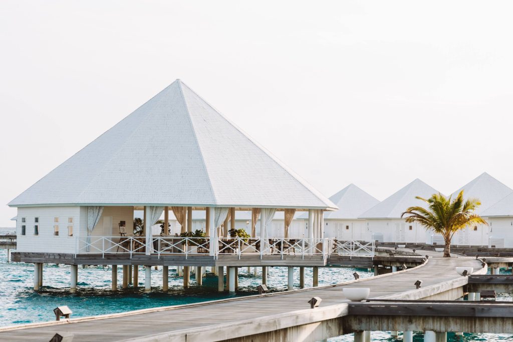 Wooden overwater bungalows with white roofs sit above turquoise ocean water, connected by a curving wooden walkway. A palm tree grows near the bungalows under a bright, clear sky.