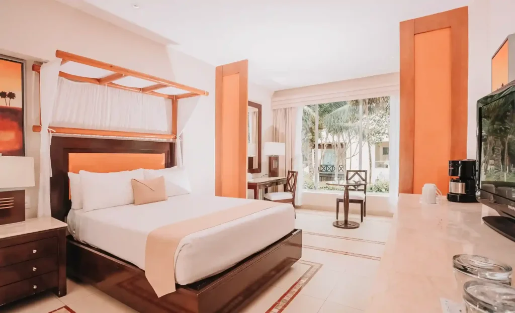 A bright, modern hotel room with a large bed, orange accents, a desk, chairs by a window, and a coffee maker on a counter. Large windows let in natural light and views of palm trees outside.