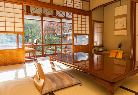 A traditional Japanese room with tatami mats, a low wooden table, cushion seating, shoji screens, and a view of a garden with autumn foliage outside the large windows. A hanging scroll and flower arrangement decorate the room.