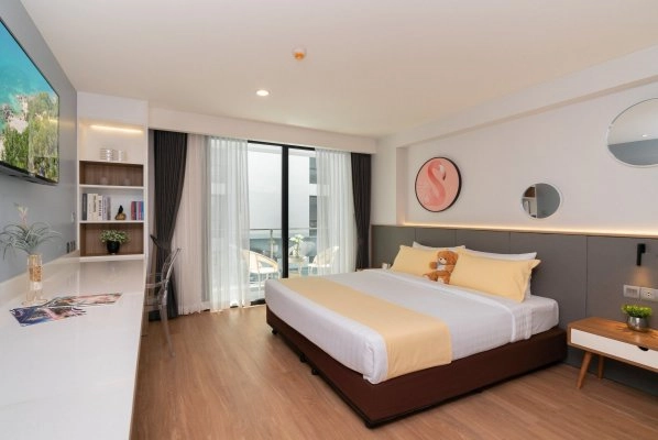 A modern hotel room with a large bed, yellow bedding, a teddy bear, a wall-mounted TV, a desk with a chair, a bookshelf, and floor-to-ceiling windows with sheer curtains leading to a balcony.