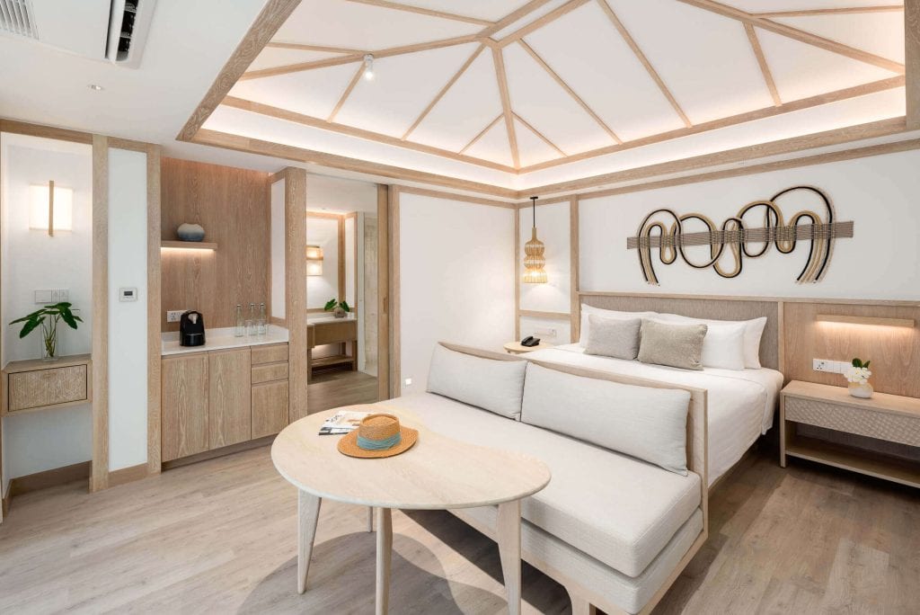A modern hotel room with a neutral color palette, featuring a bed, a cushioned bench, a small dining table with a straw hat, wall art, built-in storage, and soft natural lighting.
