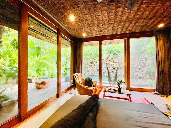 A cozy room with large glass doors overlooks a lush garden and hammock. A person relaxes on a bed, feet visible, facing the outdoor greenery. Warm lighting and natural wood accents create a tranquil, inviting atmosphere.