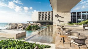 A modern beachfront resort with lounge chairs, umbrellas, and a pool beside the ocean. The stone building features two dolphin sculptures on its wall, and palm trees and greenery decorate the sunny terrace.