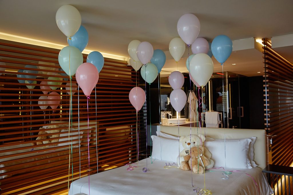 A bed in a modern room is decorated with colorful balloons and two teddy bears. The bed has white linens, and the background features wooden slats and contemporary decor.