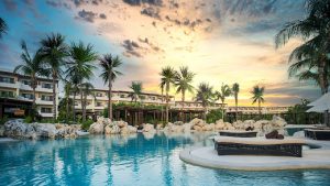 A luxurious resort pool surrounded by palm trees and lounge chairs, with a modern hotel building in the background under a colorful sunset sky.