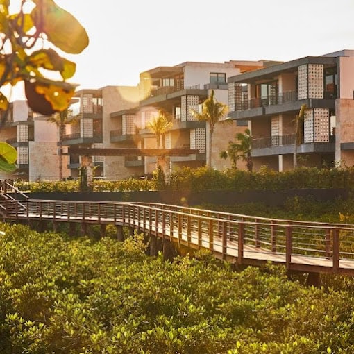 Modern resort buildings with large balconies are shown at sunset. A wooden boardwalk leads through lush green vegetation toward the buildings, with sunlight streaming through palm trees and leaves in the foreground.
