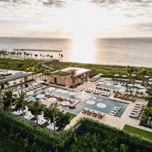 A luxurious beachfront resort with multiple pools, lounge chairs, umbrellas, palm trees, and manicured greenery, overlooking the ocean at sunset.