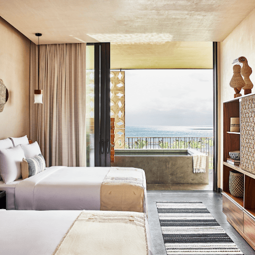 A modern hotel room with two double beds, neutral decor, woven accents, an open shelf, and large glass doors leading to a balcony with a bathtub and a view of the ocean.
