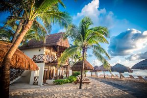 A tropical beach resort with thatched-roof buildings and palm trees, lounge chairs on the sandy shore, and a bright blue sky with scattered clouds over the ocean.