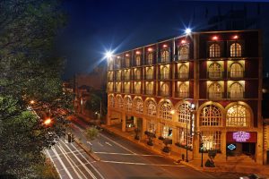 Nighttime view of a well-lit, multi-story building with large arched windows and glowing signs, located along a tree-lined street with light trails from passing vehicles.