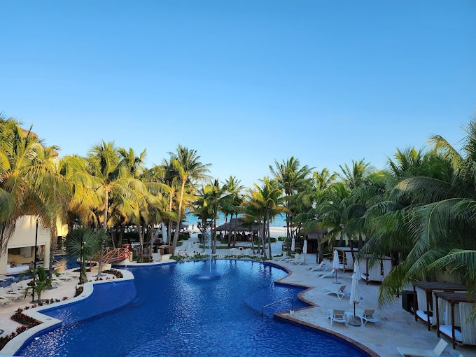 A large outdoor swimming pool surrounded by palm trees, lounge chairs, and cabanas under a clear blue sky, with the ocean visible in the background.
