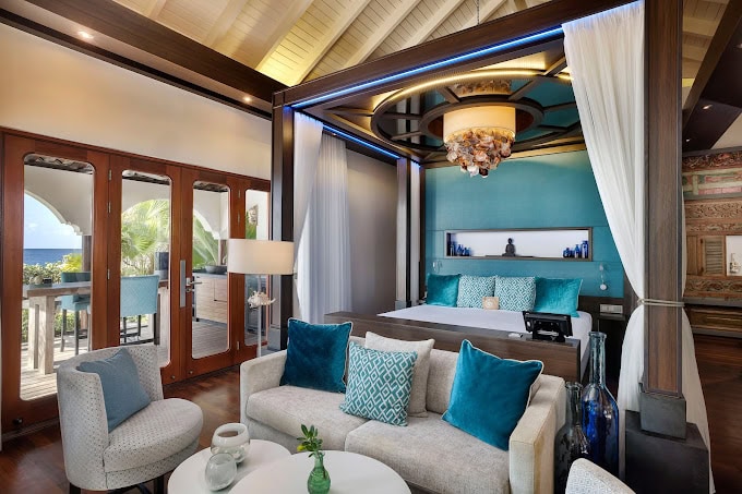 A luxurious hotel suite with a canopy bed, teal and blue decor, a plush sofa, modern chairs, and large windows letting in natural light, with views of greenery outside.