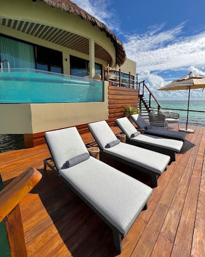 Four lounge chairs on a wooden deck beside a pool and stairs leading to clear turquoise water, with a thatched-roof villa and umbrella under a sunny, blue sky with scattered clouds.