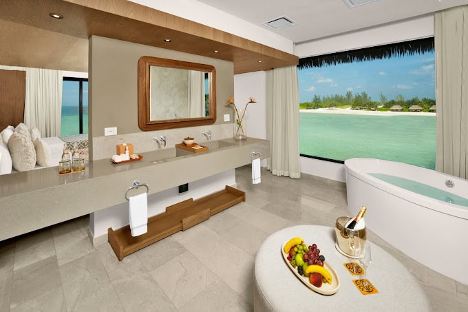 A luxurious bathroom with a double sink, large mirror, freestanding bathtub, fruit platter, and champagne, featuring a large window with a view of turquoise water and overwater bungalows.